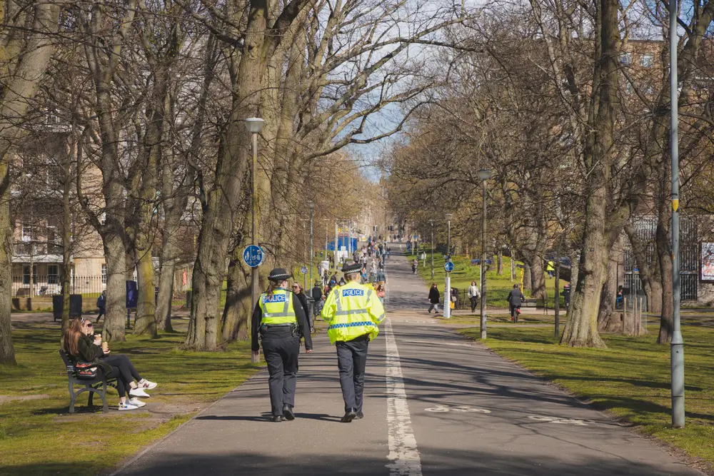 Two Police Scotland officers on foot patrol through a tree-lined urban park wearing high-visibility uniforms.