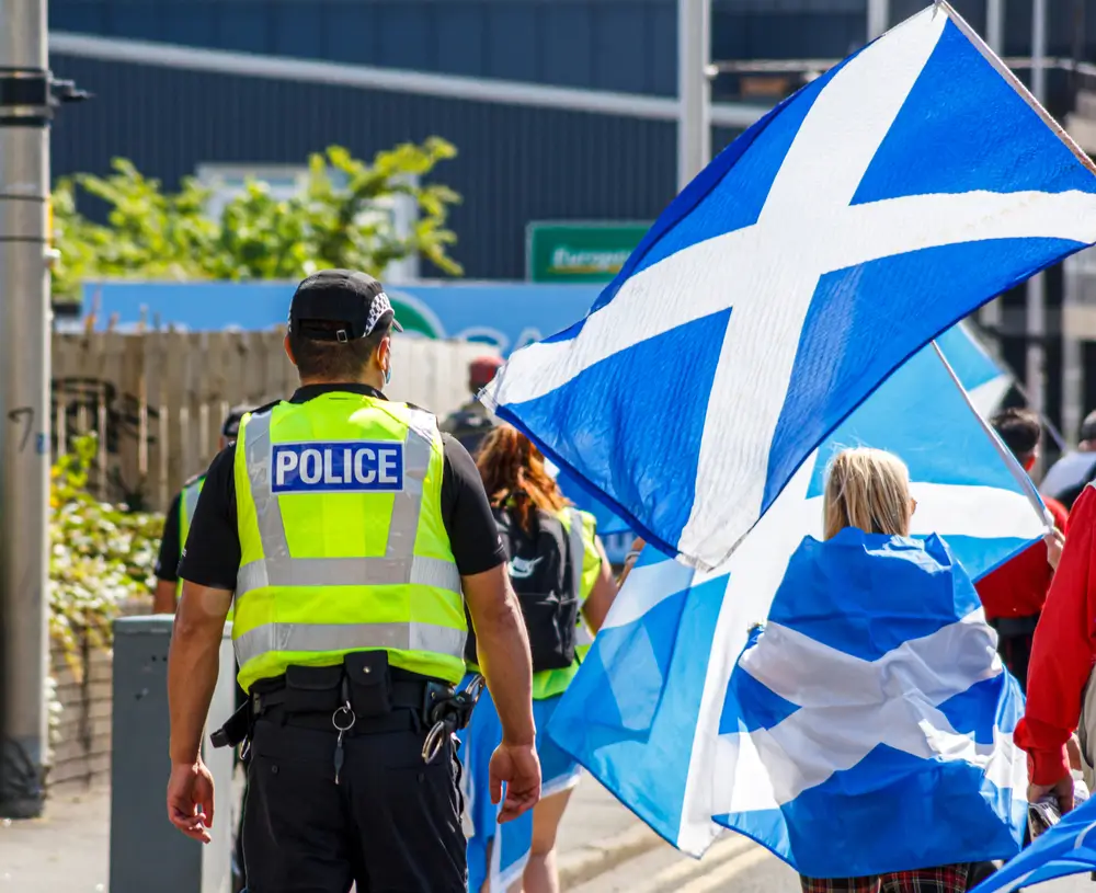 Scottish police officer in a high-visibility vest monitors a crowd waving Saltire flags during a public gathering.
