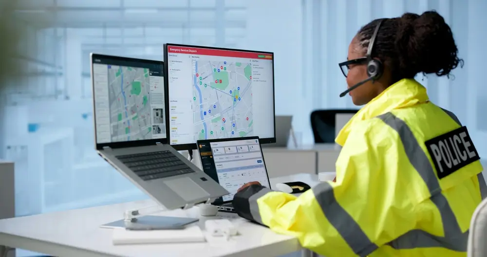 Police emergency call handler using a headset to monitor dispatch screens and maps in a modern control room.